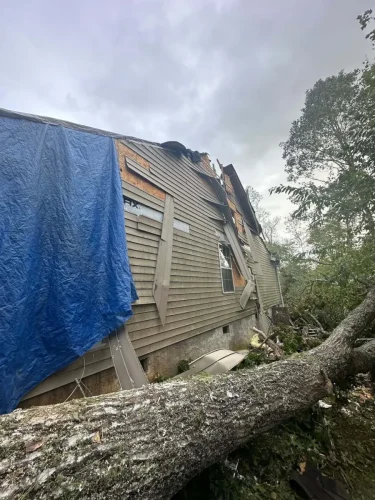 Storm damage to home exterior with fallen tree and blue tarp on roof.
