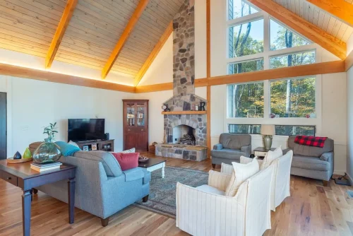 Living room with a stone fireplace, vaulted wood ceiling, exposed beams, and large windows overlooking the woods.