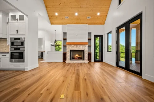 Open living area featuring wide-plank wood flooring, a wood-paneled vaulted ceiling, white fireplace, and black-framed glass doors overlooking the lake.
