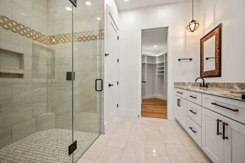 Bathroom featuring a glass-enclosed tiled shower, white double-sink vanity, dark hardware, and light-colored tile flooring.