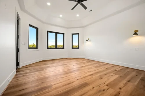 Spacious bedroom featuring wide-plank wood flooring, a tray ceiling with a black ceiling fan, recessed lighting, and black-framed windows overlooking the surrounding landscape.