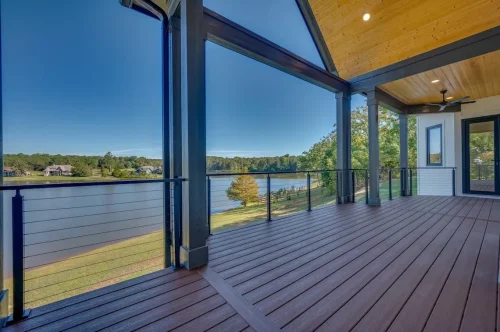 Spacious covered deck overlooking a lake, featuring wood-look decking, exposed beams, a wood-paneled ceiling, and cable railings.