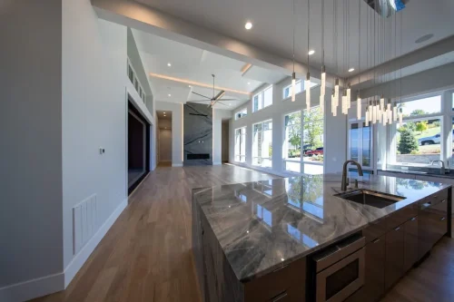 Open-concept interior with a large stone-topped kitchen island, modern pendant lighting, wood flooring, and a fireplace beyond.