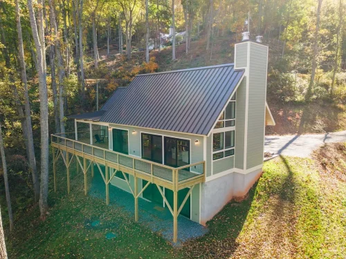 Elevated exterior view of a green mountain home with a metal roof and wraparound deck in a wooded setting.