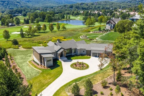 Drone shot of the property showing the home, landscaped yard, and nearby golf course with pond and mountains.