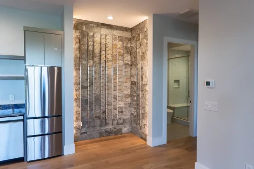 Built-in wine storage display set against a textured stone wall beside a stainless steel refrigerator and hallway.