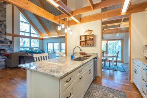 Kitchen with a large stone countertop island, white cabinetry, exposed wood beams, and views into the living and dining areas.