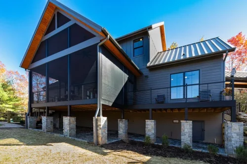 Rear view of the mountain home featuring a large screened porch, elevated deck, metal roof, and stone column bases.