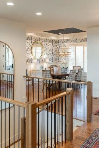 Elegant dining room with blue and white wallpaper, gold chandelier, round wood table, and upholstered chairs, framed by white plantation shutters.