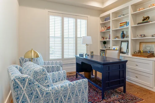 Traditional office with navy desk, built-in shelving, patterned blue chairs, and natural light from tall windows with plantation shutters.