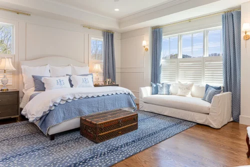 Spacious primary bedroom decorated in blue and white with a tufted headboard, patterned rug, and a white settee beneath large windows.