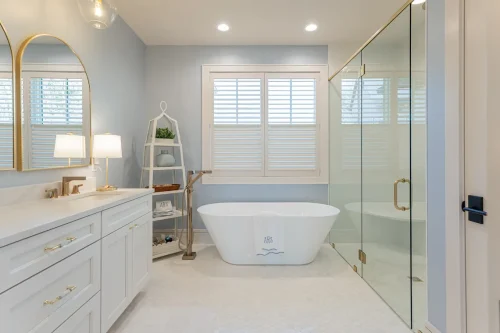Luxurious bathroom with a freestanding tub, glass shower, double vanity with gold mirrors, and light blue walls.