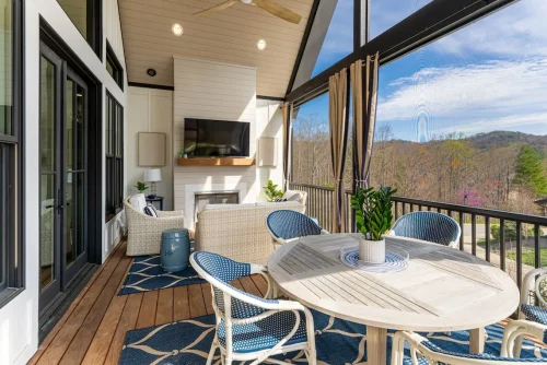 Screened-in porch with vaulted ceiling, outdoor fireplace, mounted TV, wicker seating, and dining table overlooking wooded views.
