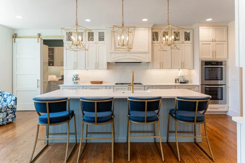 Bright white kitchen with quartz countertops, gold pendant lighting, and navy bar stools, featuring a large island and double wall ovens.