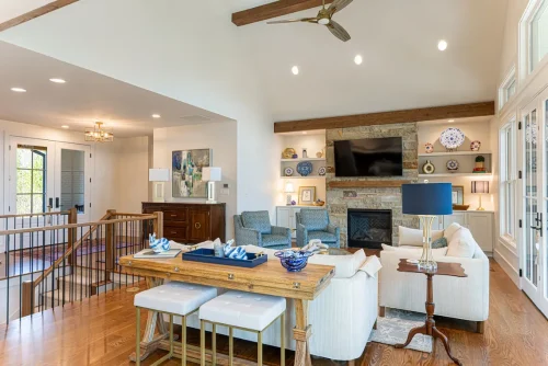 Cozy living room with vaulted ceilings, stone fireplace, built-in shelving with blue and white ceramics, and neutral seating accented with patterned chairs.