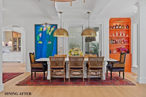 Renovated dining room with a white dining table, woven chairs, brass pendant lights, colorful artwork, and bright orange built-in shelving.