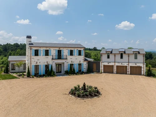 Stately white stone home with blue shutters, wood garage doors, circular driveway, and central fountain set against rolling mountain views.