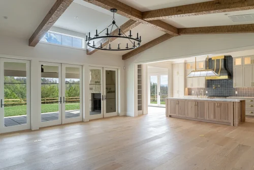 Open-concept layout with rustic wood beams, black iron chandelier, and sightline from living area to kitchen with custom range hood.