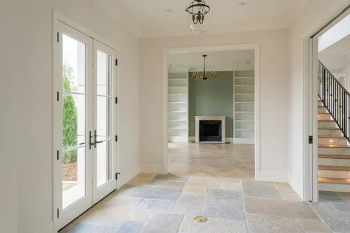 Elegant entry foyer with stone tile flooring, glass French doors, and view into formal living room with green accent wall, built-in shelving, and fireplace.