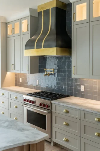 Chef’s kitchen with custom black and gold range hood, marble counters, gray cabinetry with brass hardware, and tile backsplash.
