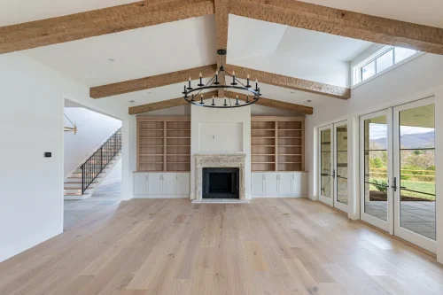 Spacious living area with exposed wood beams, stone fireplace, built-in shelving, and oversized windows opening to mountain views.