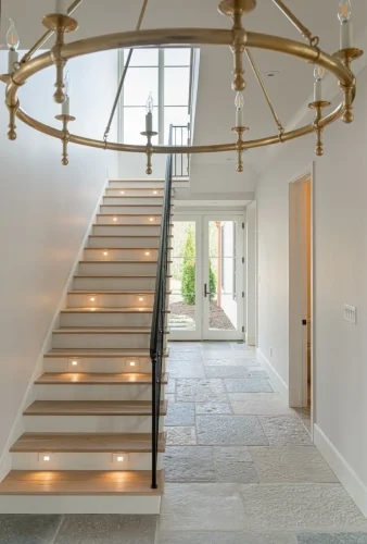 Grand staircase with stone flooring, black iron railing, lighted steps, and large gold chandelier in two-story foyer.