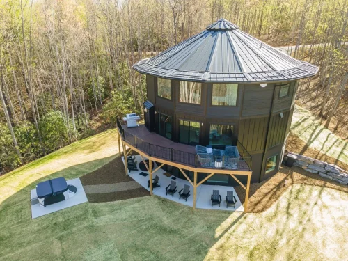 Striking round home with a dark exterior, large deck, and lower patio with seating, set on a hillside among trees.