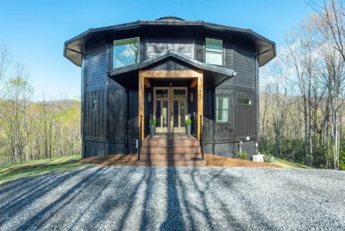 Round mountain home with black siding, wood-trimmed entry, and large windows reflecting the forest.