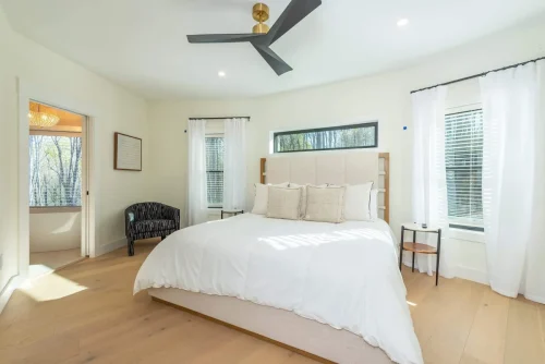 Airy bedroom with a king bed, modern ceiling fan, white curtains, and large windows filling the space with natural light.