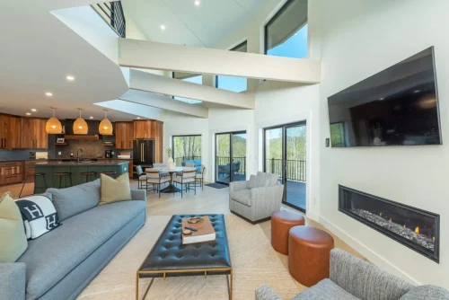 Modern living space with vaulted ceilings, wall-mounted TV above sleek fireplace, and open flow to kitchen and dining.