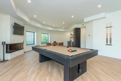 Spacious entertainment room with pool table, TV, corner fireplace, and bar seating, framed by large windows.