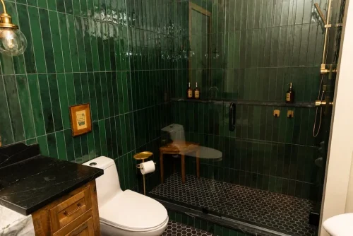Bathroom with deep green tile, glass-enclosed walk-in shower, wood vanity, and black stone countertop.