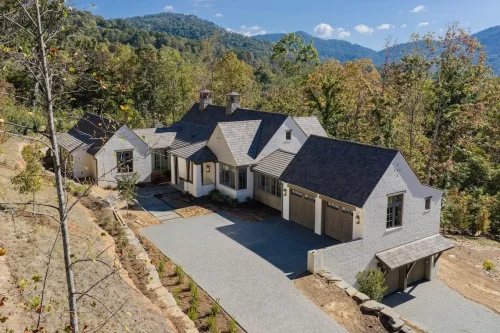 Aerial view of a mountain home with white brick siding, dark gabled rooflines, wood garage doors, and a paved driveway surrounded by trees.