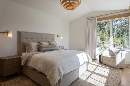 Bedroom featuring a tufted upholstered bed, neutral furnishings, wood nightstands, and large windows overlooking the trees.