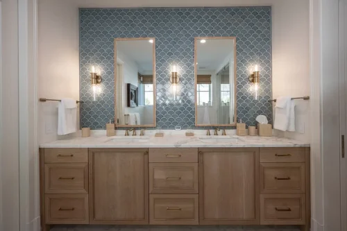Double vanity with natural wood cabinetry, marble countertops, brass fixtures, and a blue scalloped tile accent wall.