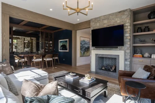 Living area featuring a stone fireplace, built-in shelving, neutral furnishings, and an adjoining bar with dark cabinetry and wood accents.
