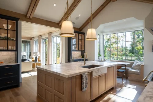 Spacious kitchen featuring a large marble island, natural wood cabinetry, exposed ceiling beams, pendant lighting, and a breakfast nook surrounded by windows.