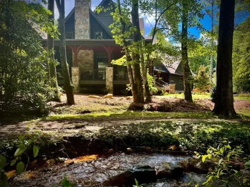 Mountain home with stone chimney and dark wood siding, surrounded by tall trees with a creek in the foreground.