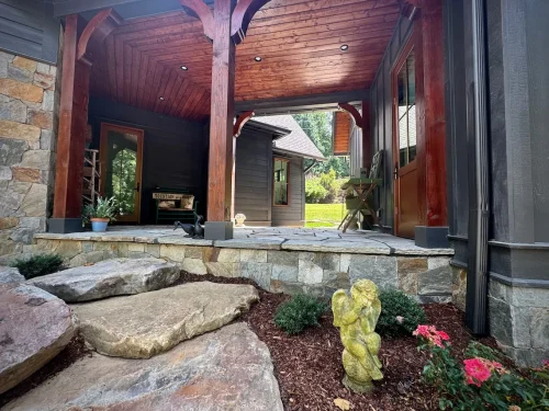 Covered porch entry with stone steps, wood beams, and landscaping featuring flowers and a small angel statue.