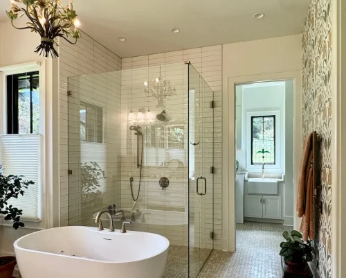 Bright bathroom with freestanding tub, glass shower, floral wallpaper accent, and natural light from windows.