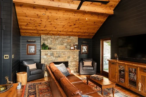 Living room with dark walls, wood-paneled ceiling, stone fireplace, leather seating, and hardwood floors.