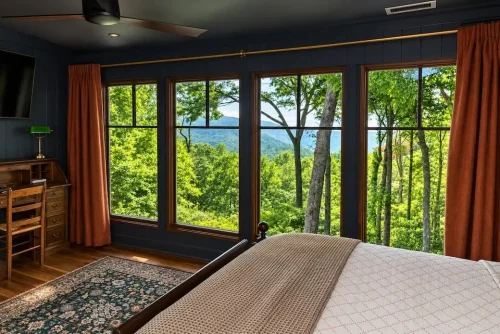 Bedroom with dark walls, hardwood floors, wood furniture, and large windows overlooking wooded mountain views.