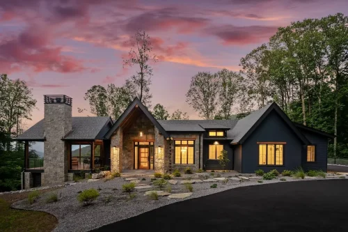 Front exterior of a contemporary mountain home featuring dark siding, natural stone, warm wood accents, and a large stone chimney at sunset.