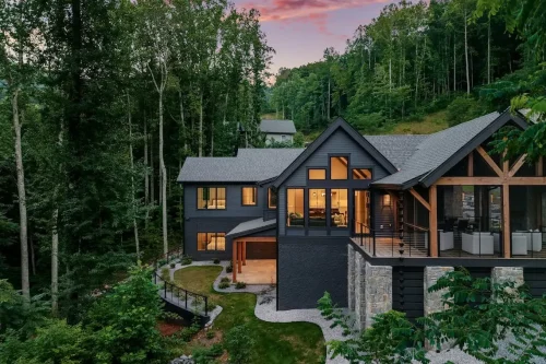 Rear exterior of a contemporary mountain home surrounded by woods, featuring dark siding, large windows, stone accents, and a covered outdoor living area.