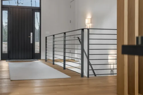 Minimalist foyer with wide plank wood floors, black front door, and sleek horizontal metal stair railing, accented by soft wall lighting.