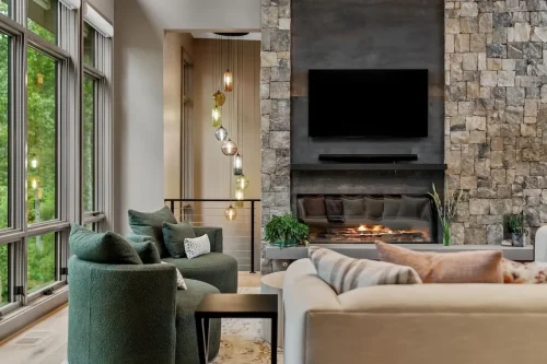 Living area with a floor-to-ceiling stone fireplace, modern firebox, and expansive windows overlooking the wooded setting.