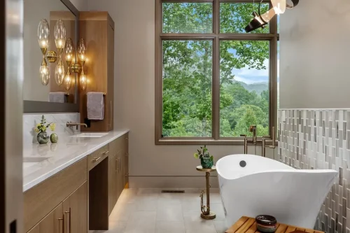 Primary bathroom with a freestanding soaking tub, wood cabinetry, modern lighting, and a large window framing the mountain view.