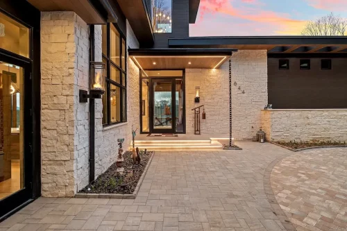Covered entry with light stone walls, black-framed windows and doors, illuminated steps, and a paved courtyard.