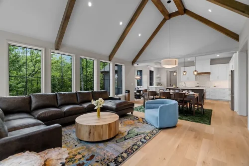 Open-concept great room with vaulted ceilings, exposed wood beams, large windows, hardwood floors, and views into the dining area and kitchen.