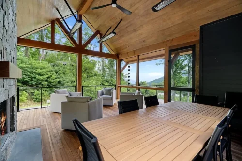 Screened outdoor living and dining area with a wood-paneled vaulted ceiling, exposed beams, stone fireplace, and wooded mountain views